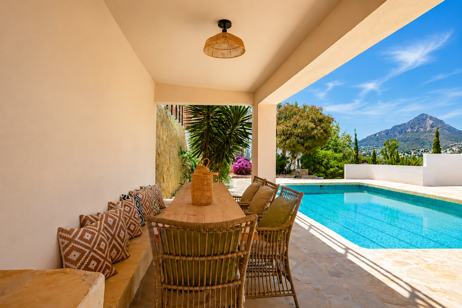 Espace repas extérieur couvert avec table en bois et chaises en osier, bordant la piscine. Vues sur le Montgó et végétation méditerranéenne sur un sol en pierre.