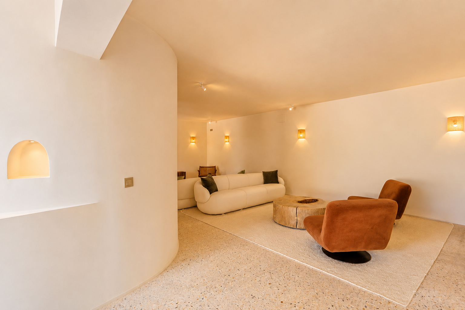 Espace salon contemporain avec canapé incurvé blanc cassé, deux fauteuils couleur rouille et table basse en bois rustique sur terrazzo clair. Des appliques murales tissées diffusent une lumière chaude.