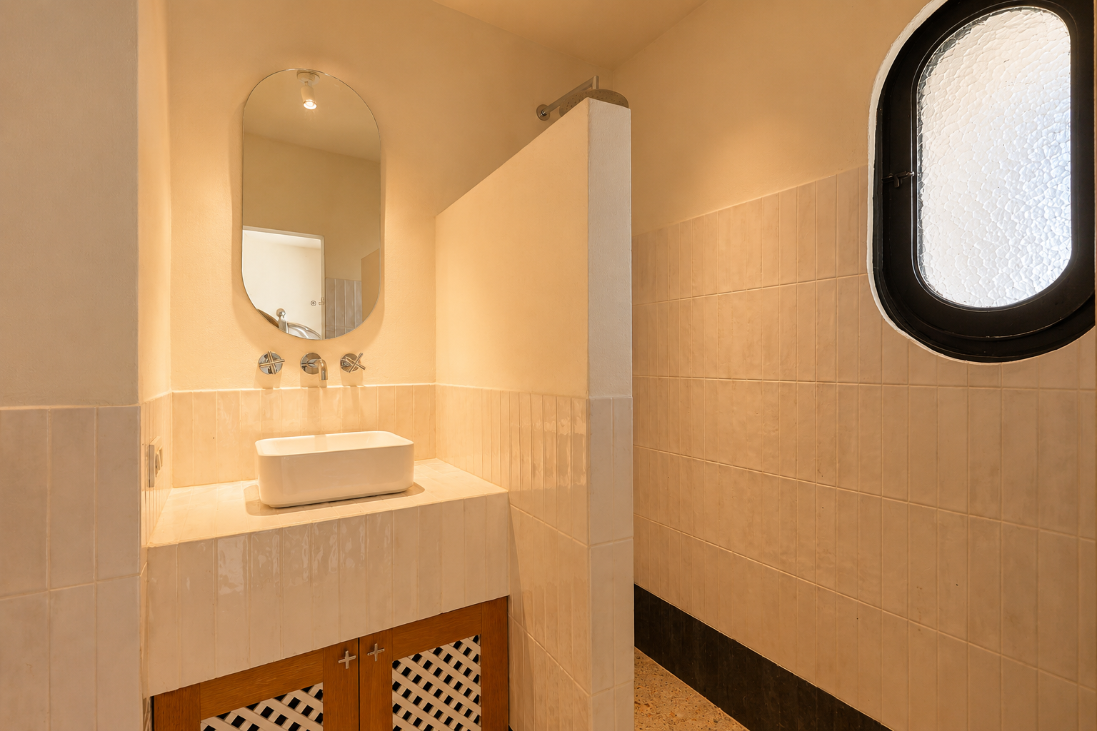 Meuble de salle de bain avec vasque rectangulaire, robinetterie murale chromée et miroir ovale. Zone de douche attenante avec fenêtre ovale dépolie et murs carrelés clairs.