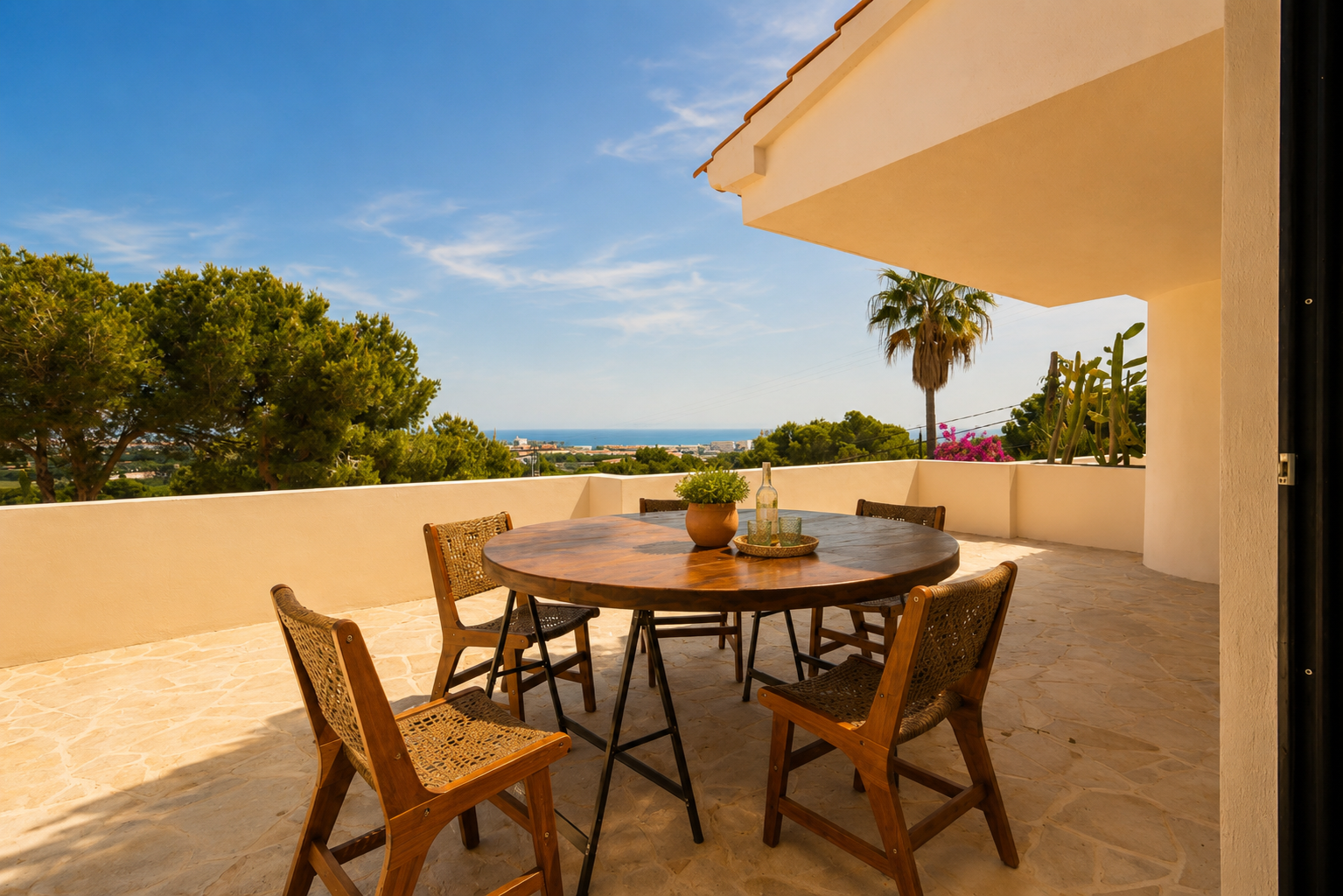 Terrasse extérieure avec table ronde en bois et chaises tressées sur dalles, offrant une vue panoramique sur la mer et la côte.