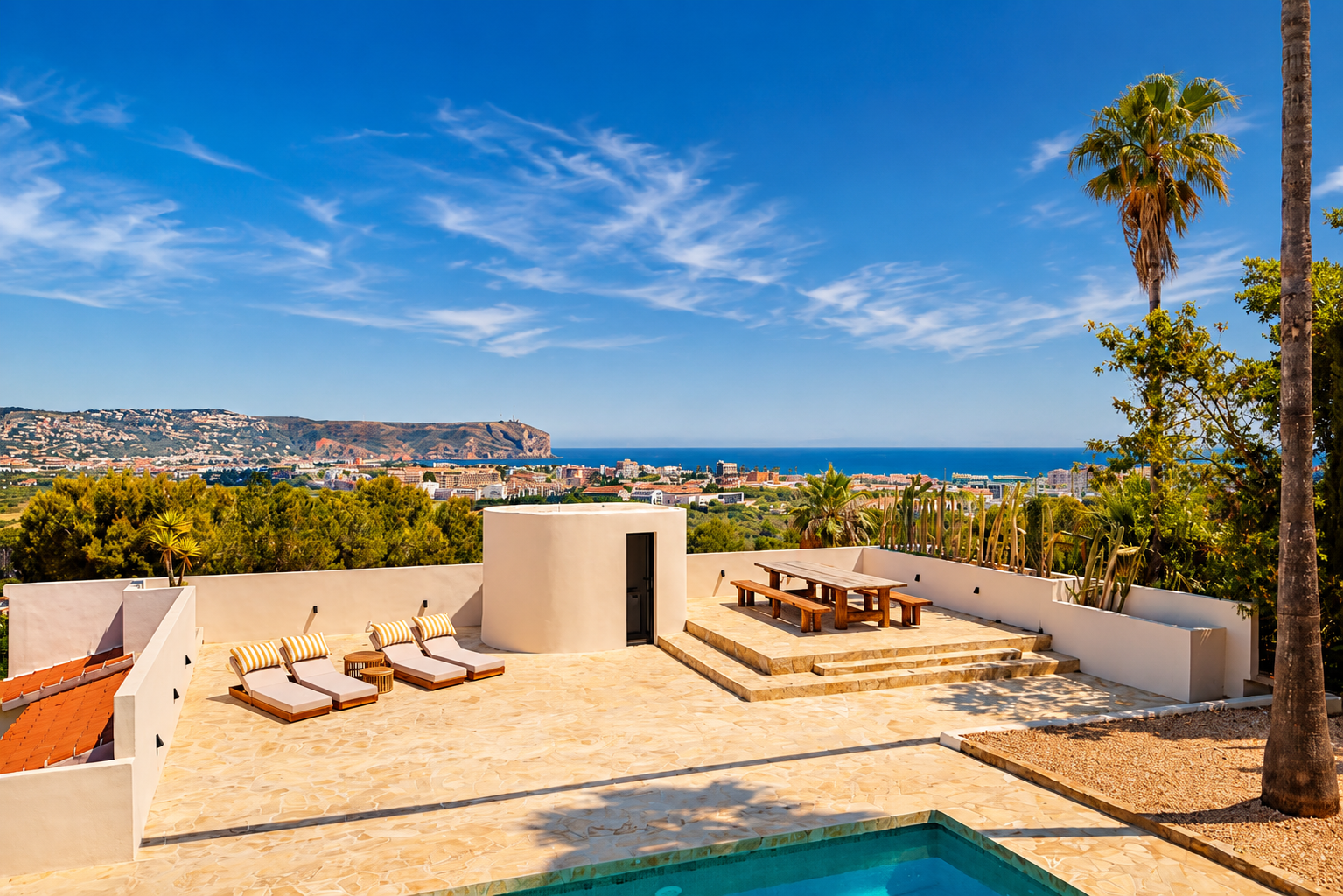 Vaste terrasse en pierre avec piscine, chaises longues et coin repas en bois surélevé, offrant une vue panoramique sur la baie de Jávea et la côte sous un ciel clair.