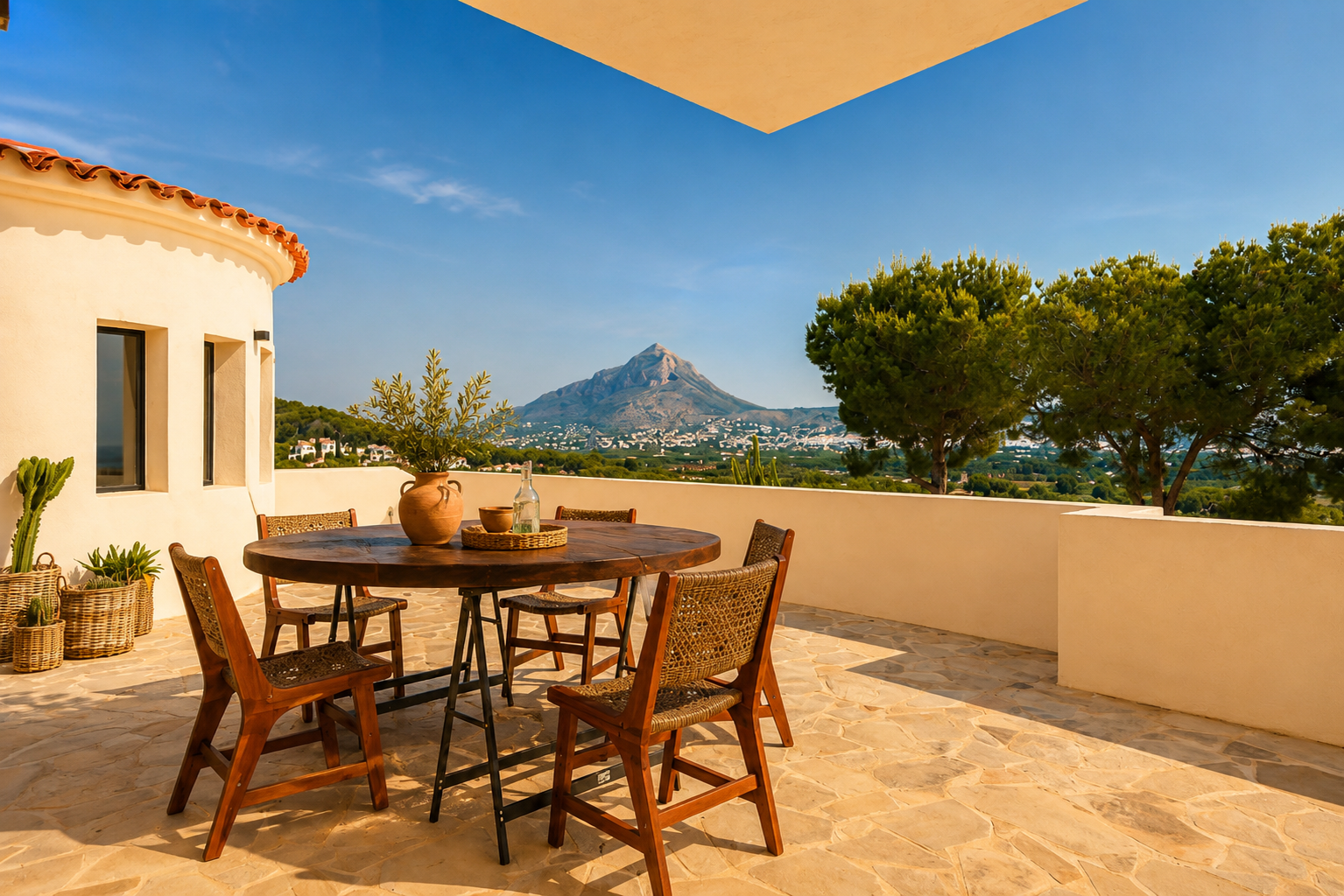 Vaste terrasse extérieure avec sol en dalles irrégulières et mobilier de repas en bois rustique. Vue panoramique sur le Montgó et la vallée sous un ciel clair.