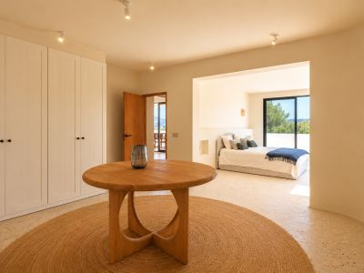 Cet espace de transition lumineux comprend une table ronde en bois sur un tapis en jute, flanquée d'armoires blanches intégrées. Il mène à une chambre avec vue sur la verdure.