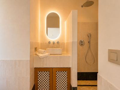 Cette salle de bain élégante présente une vasque, un miroir ovale rétroéclairé et un meuble en bois ajouré. La douche attenante est équipée d'une pomme de douche pluie.