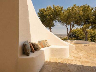 Cette terrasse extérieure offre un banc en stuc intégré aux courbes douces, agrémenté de coussins bohèmes, sur un patio en dalles irrégulières. Des pins et une vue lointaine sur le paysage sont visibles.