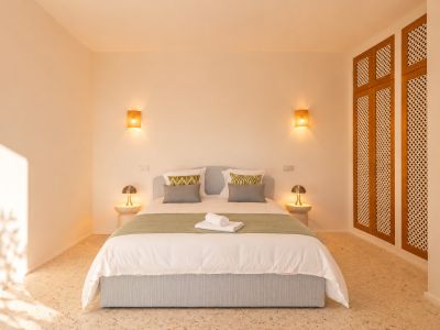 Chambre avec lit capitonné bleu clair, sol en terrazzo et éclairage chaleureux d'appliques murales tressées. Une armoire en bois à persiennes est visible à droite.
