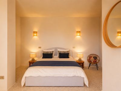 Chambre avec lit double, tables de chevet en bois et lampes de table en céramique. Des appliques murales en raphia et un miroir rond en bois ornent l'espace. Sol en terrazzo.
