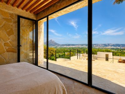 Chambre avec murs en pierre naturelle et poutres en bois, s'ouvrant sur une terrasse avec vues panoramiques sur le Montgó et la baie de Jávea.