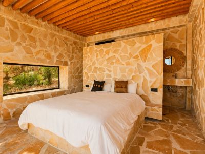 Chambre avec murs et sol en pierre naturelle irrégulière, plafond à poutres en bois et fenêtre horizontale sur la végétation. Un lavabo intégré avec miroir en rotin est présent.