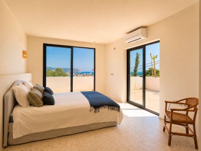 Chambre lumineuse avec sol en terrazzo clair et grandes portes coulissantes vitrées menant à une terrasse privée. Offre des vues étendues sur la mer et la côte lointaine.