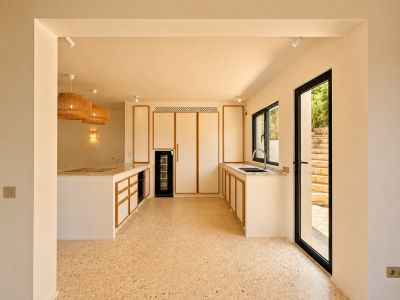 Cuisine moderne avec des armoires bordées de bois clair, un sol terrazzo et un grand îlot avec table de cuisson intégrée. Des suspensions en rotin éclairent la pièce.
