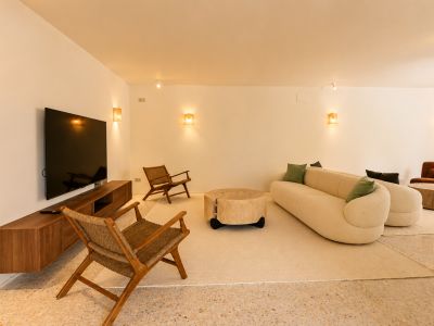 Espace de vie contemporain avec canapé incurvé crème, table basse rustique en bois et fauteuils en osier sur sol en terrazzo, éclairé par des appliques murales chaleureuses.
