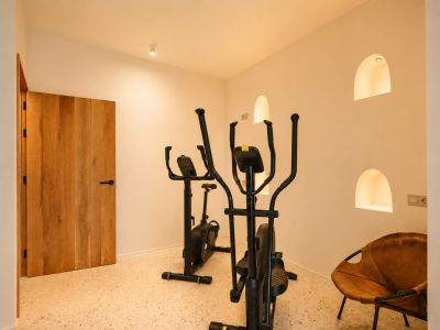 Espace fitness dédié avec vélo elliptique et vélo d'appartement sur sol en terrazzo, agrémenté de niches murales arquées éclairées et d'une porte en bois.