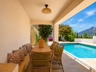 Espace repas extérieur couvert avec table en bois et chaises en osier, bordant la piscine. Vues sur le Montgó et végétation méditerranéenne sur un sol en pierre.