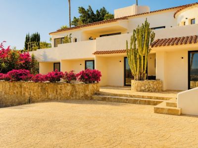 Façade crème de la villa avec tuiles en terre cuite, ornée de bougainvilliers fuchsia et d'un grand cactus, menant à une allée pavée et des marches en pierre.