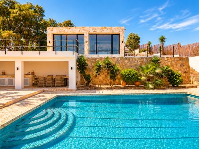 Grande piscine extérieure avec marches incurvées, bordée d'une terrasse pavée menant à un espace repas couvert et cuisine extérieure. Au-dessus, un bâtiment en pierre avec balcon et portes vitrées.