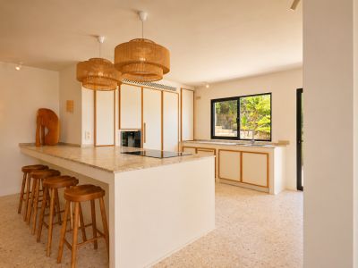 Îlot de cuisine moderne avec plaque de cuisson intégrée, comptoir clair moucheté et cinq tabourets en bois. Des suspensions tressées éclairent l'espace, offrant une vue sur la verdure luxuriante.