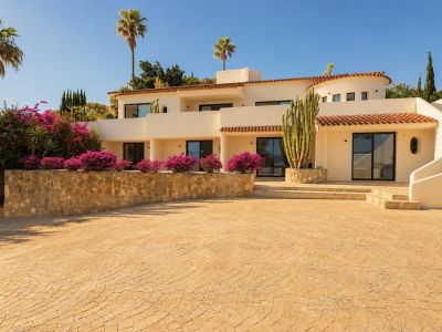 La façade crème de la villa, les tuiles de terre cuite et les grandes portes vitrées, encadrées par des bougainvilliers éclatants, un grand cactus et des palmiers sous un ciel bleu. Une allée pavée mène à l'entrée.