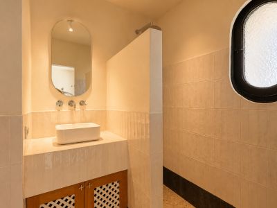 Meuble de salle de bain avec vasque rectangulaire, robinetterie murale chromée et miroir ovale. Zone de douche attenante avec fenêtre ovale dépolie et murs carrelés clairs.