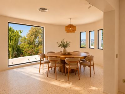Salle à manger lumineuse avec table en bois organique pour huit personnes et sol en terrazzo. De grandes fenêtres incurvées encadrent la végétation luxuriante et des vues lointaines.