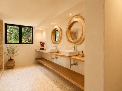 Salle de bain avec double vasque en bois, lavabos rectangulaires blancs et grands miroirs ronds aux cadres en bois naturel. Une fenêtre donne sur la verdure, complétant le sol en terrazzo clair.