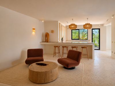 Salon ouvert avec deux fauteuils rouille, une table basse rustique en bois et sol en terrazzo clair. Un bar avec tabourets et suspensions tressées offre une vue sur la verdure extérieure.