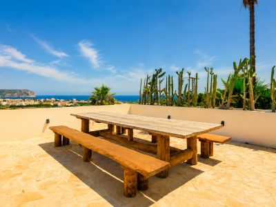 Terrasse extérieure avec table et bancs en bois rustique, sol en pierre claire, offrant des vues panoramiques sur la mer et la ville, bordée de cactus.