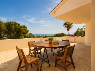 Terrasse extérieure avec table ronde en bois et chaises tressées sur dalles, offrant une vue panoramique sur la mer et la côte.