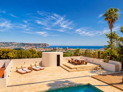 Vaste terrasse en pierre avec piscine, chaises longues et coin repas en bois surélevé, offrant une vue panoramique sur la baie de Jávea et la côte sous un ciel clair.