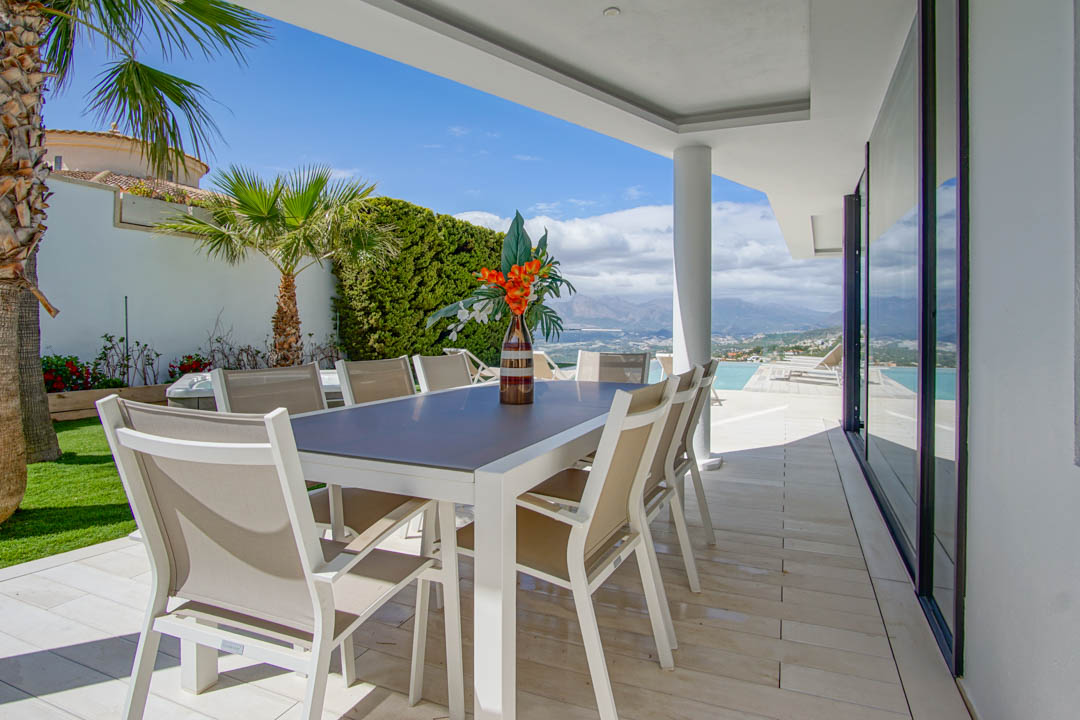 Cette terrasse repas extérieure couverte dispose d'une table blanche moderne et de huit chaises beiges sur carrelage clair. Elle offre des vues étendues sur les montagnes et la mer, à côté de la piscine à débordement.