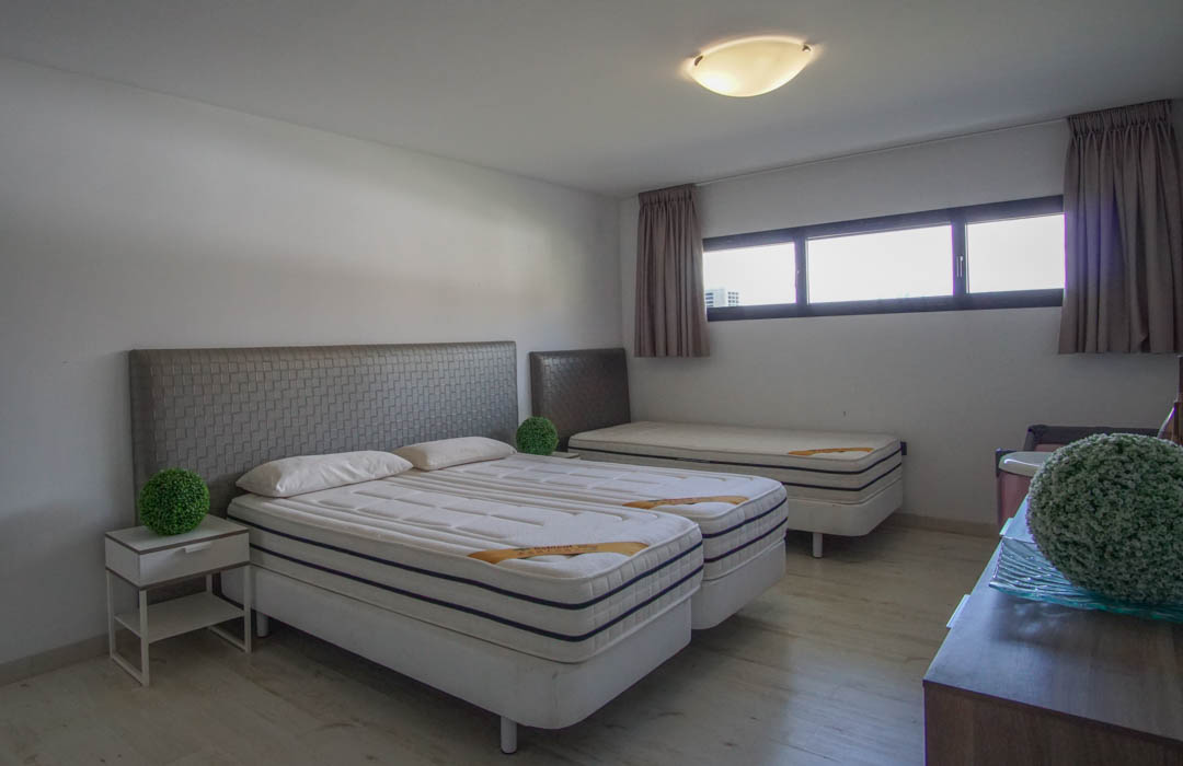 Chambre à coucher avec deux lits simples combinés, têtes de lit grises texturées, un troisième lit simple à l'arrière, parquet clair et fenêtres horizontales.