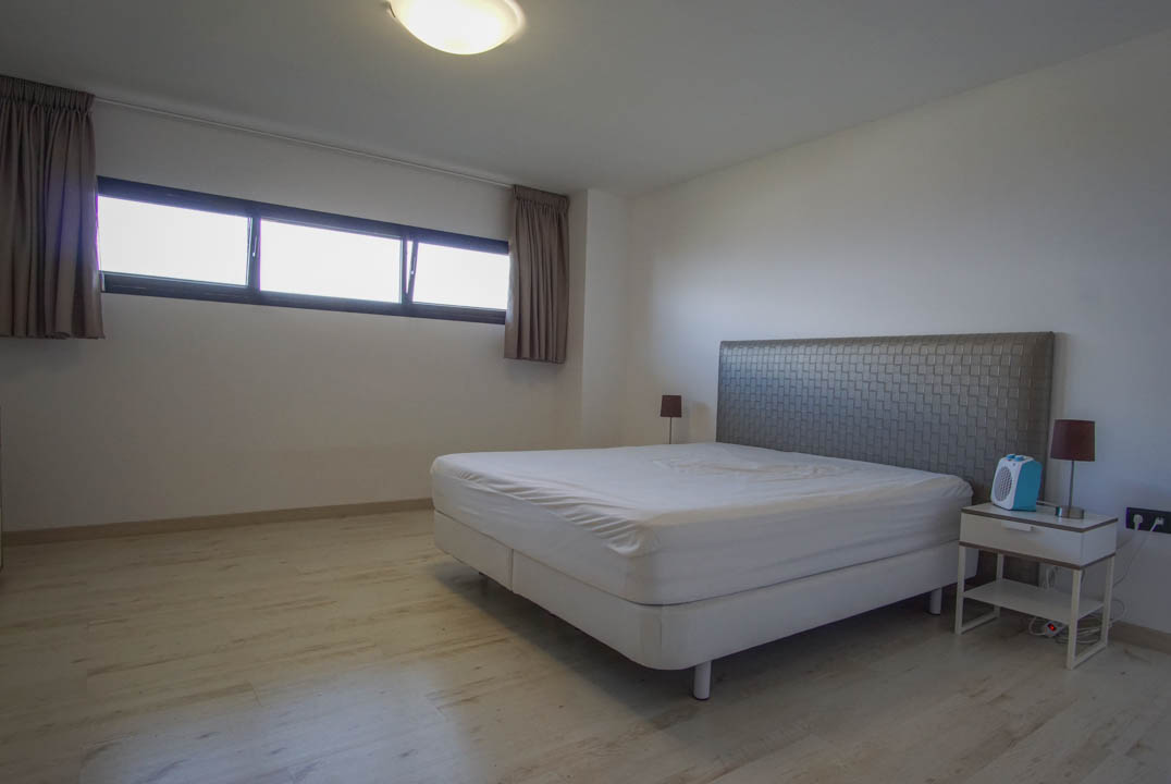 Chambre à coucher avec sol aspect bois clair et murs blancs. Comprend un lit avec tête de lit texturée, une table de chevet et une fenêtre horizontale encadrée de noir.