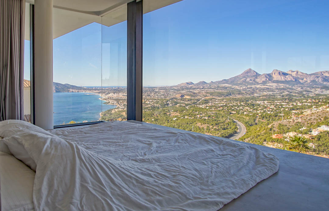 Chambre avec baies vitrées du sol au plafond, encadrant des vues panoramiques sur la mer Méditerranée, une ville côtière et les montagnes lointaines. Un lit bas avec linge blanc fait face à la vue.