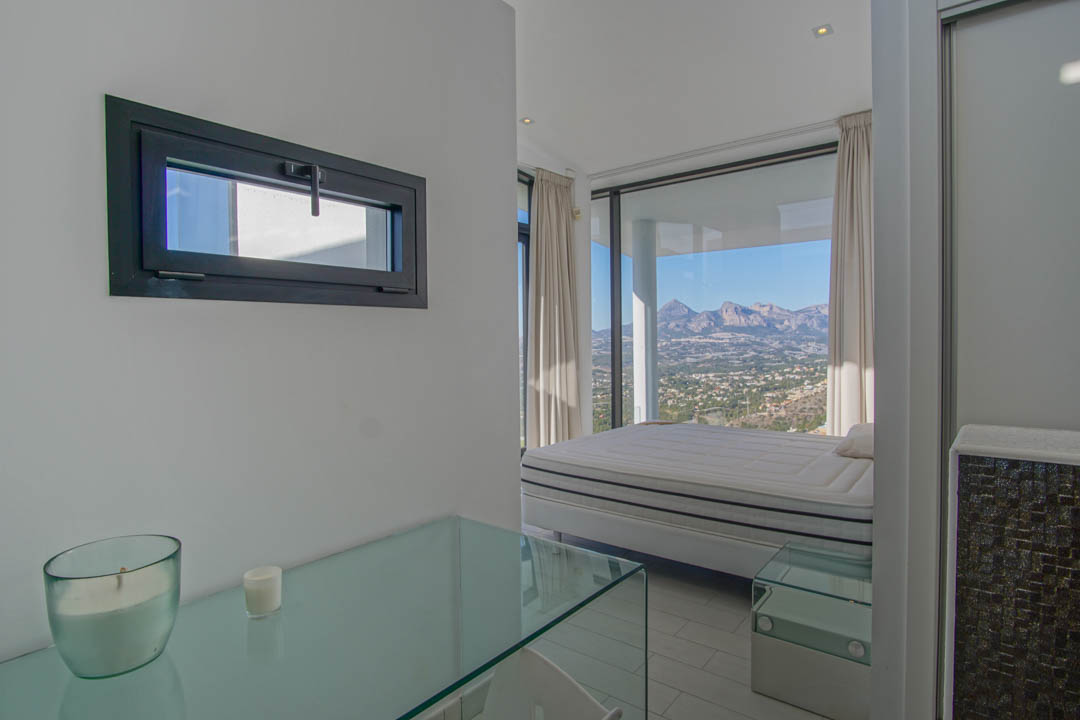Chambre lumineuse avec un lit placé devant de grandes baies vitrées, offrant une vue panoramique sur les montagnes et la vallée. Une console en verre et une fenêtre horizontale sont visibles.