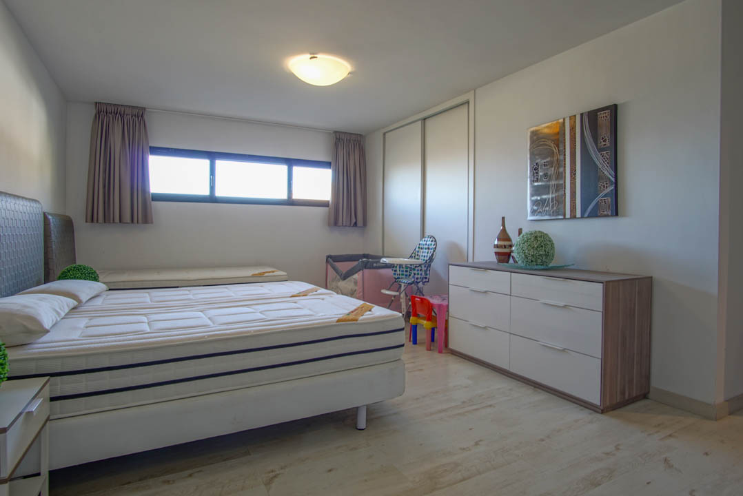 Chambre lumineuse dotée d'un lit avec deux matelas, d'un sol imitation bois clair et d'une longue fenêtre horizontale. Elle comprend une armoire coulissante blanche et une commode moderne surmontée d'une œuvre abstraite.