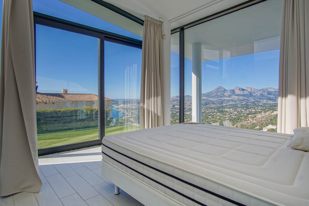 Chambre moderne avec grand lit et vastes fenêtres. Elle offre des vues panoramiques sur la mer lointaine, une pelouse verte et des montagnes escarpées sous un ciel clair.