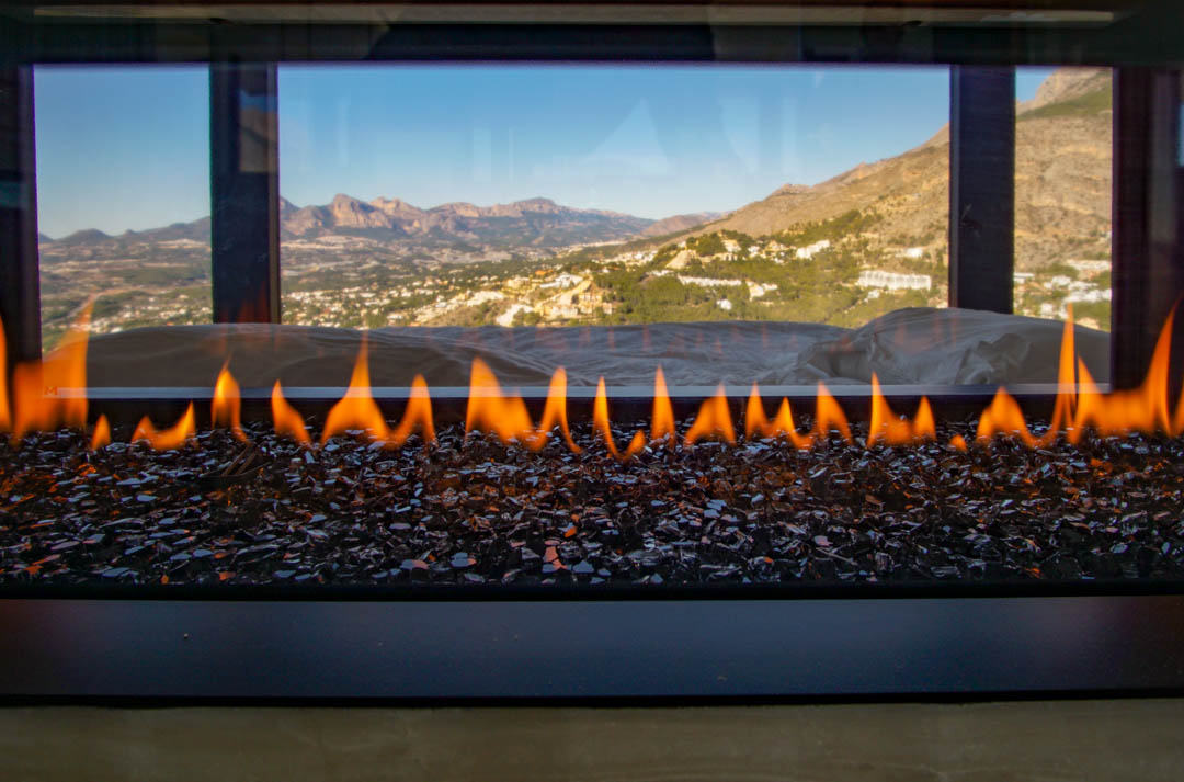 Cheminée à gaz contemporaine avec flammes sur lit de verre sombre, offrant une vue panoramique sur les montagnes et la vallée.