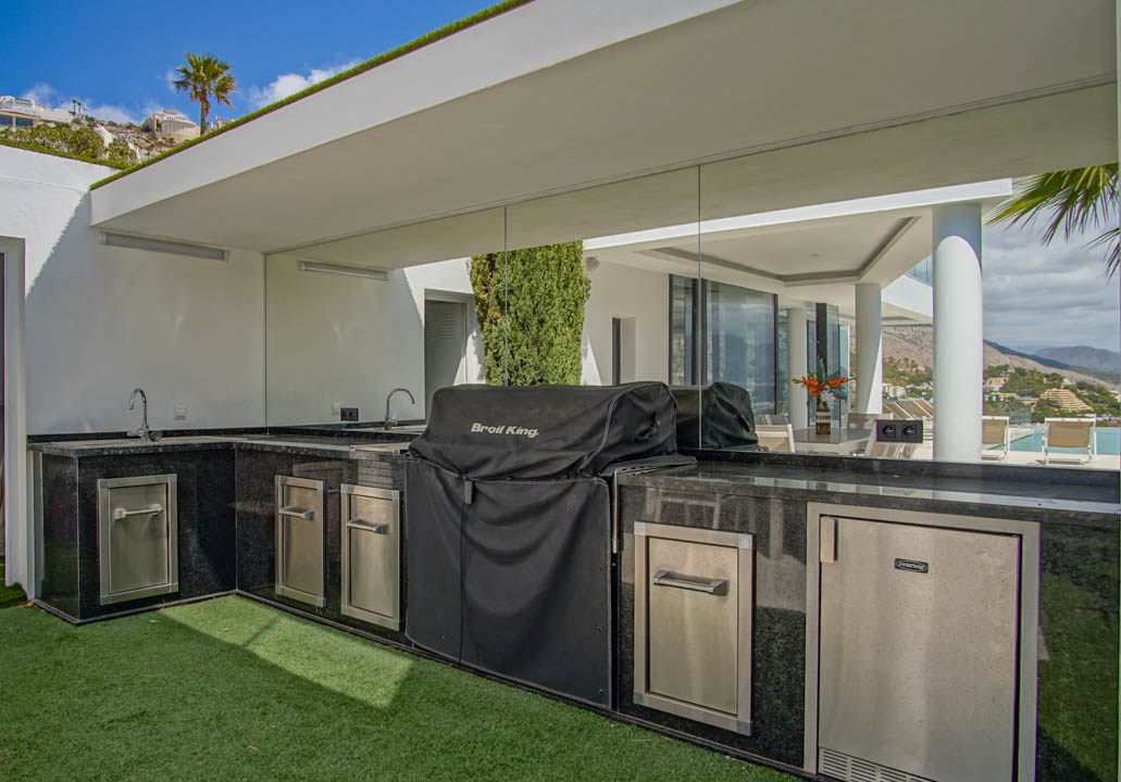 Cuisine extérieure en L avec plans de travail en granit noir, double évier, barbecue couvert et appareils en acier inoxydable, reflétant la vue sur la piscine.