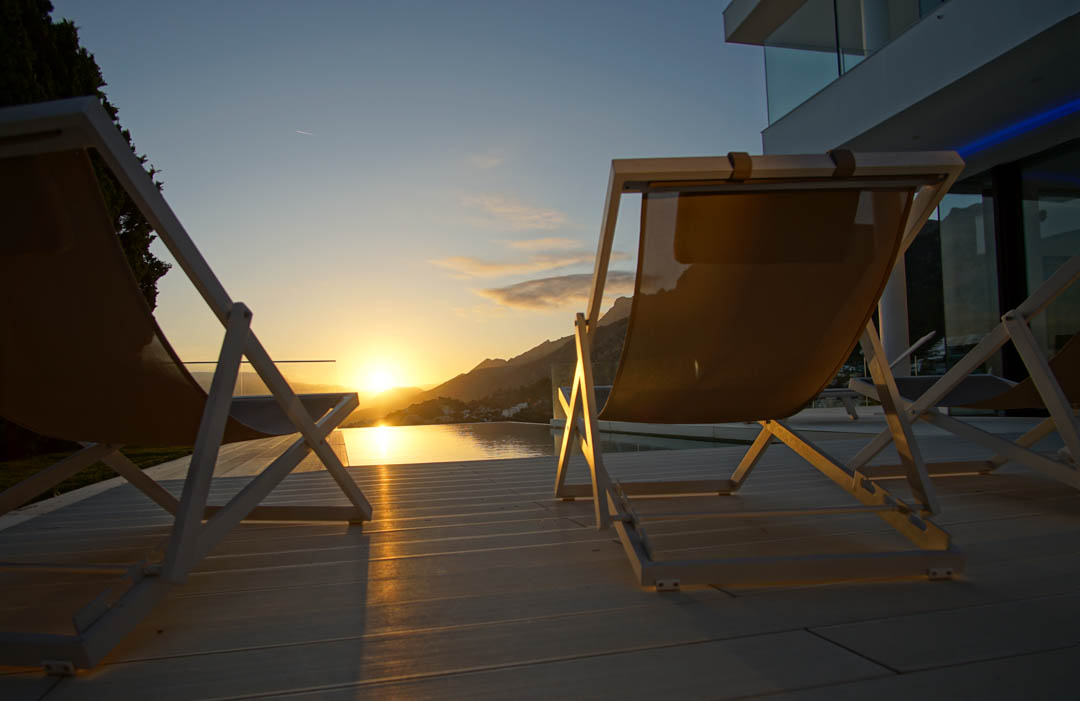 Deux chaises longues modernes sur une terrasse claire surplombent une piscine à débordement, reflétant un coucher de soleil doré sur les montagnes.