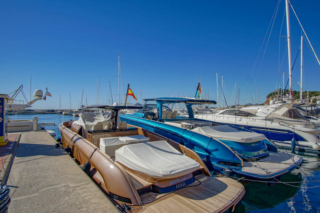 Deux vedettes modernes, l'une bronze avec pont en teck et l'autre bleu métallique, amarrées dans une marina par ciel clair. Des drapeaux espagnols ornent les bateaux, avec d'autres yachts en arrière-plan.