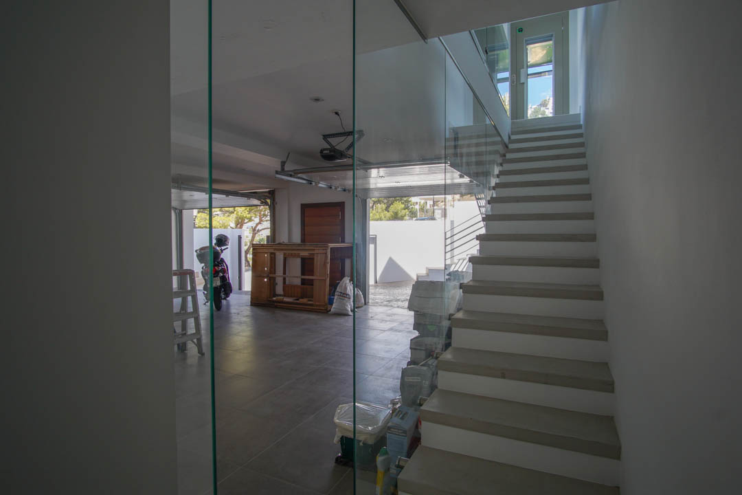 Escalier intérieur moderne avec balustrade en verre et marches claires, jouxtant un garage carrelé avec porte ouverte et lumière naturelle.