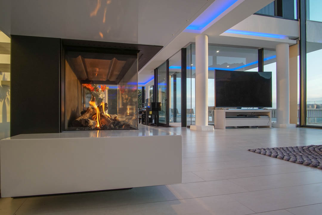Espace de vie moderne avec cheminée à gaz vitrée et base blanche. De grandes fenêtres et un éclairage d'accentuation bleu complètent le design.