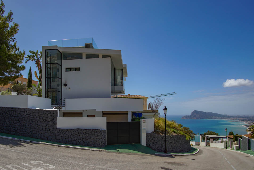 Extérieur d'une villa moderne blanche avec un ascenseur en verre et une balustrade de terrasse sur le toit. Murs de soutènement en pierre et portail automatique, avec vue panoramique sur la mer.