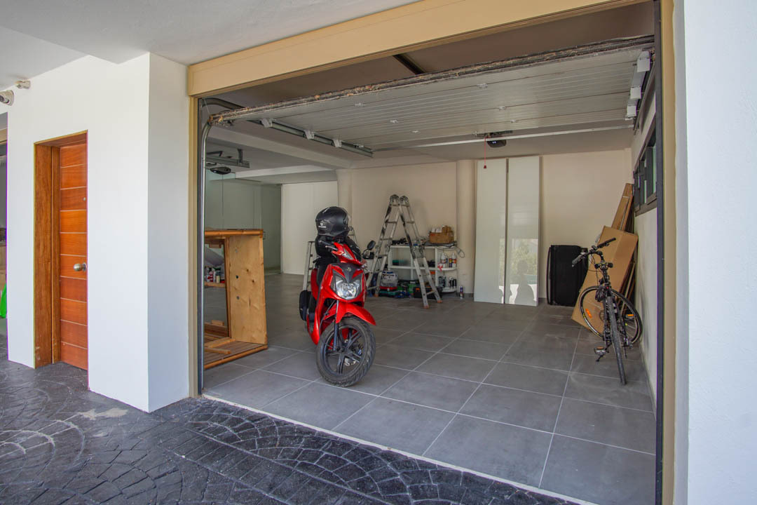 Grand garage avec carrelage gris, porte sectionnelle partiellement ouverte et porte d'entrée en bois distinctive à gauche. Une trottinette rouge et un vélo sont visibles.
