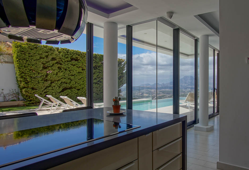 Îlot de cuisine moderne avec plan de travail sombre et plaque de cuisson intégrée. Les baies vitrées offrent une vue sur la piscine à débordement, la vallée et les montagnes.