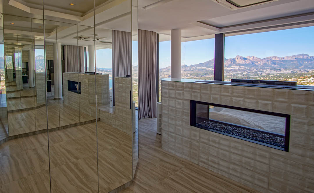 Intérieur contemporain avec cheminée à gaz transparente sur lit de galets, intégrée dans un mur texturé. Des panneaux miroir reflètent la pièce et les vues imprenables sur les montagnes et la mer.