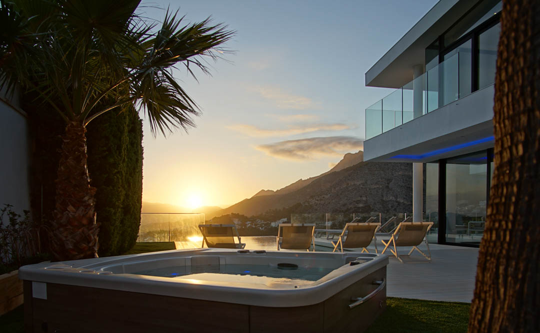 Jacuzzi extérieur avec finition bois, sur pelouse verte, devant une villa moderne avec balcons vitrés. Vue montagne et mer au coucher du soleil.