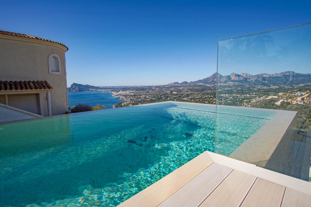 La piscine à débordement avec son eau claire, sa terrasse claire et sa barrière en verre, offre une vue panoramique sur la mer, la côte et les montagnes lointaines.