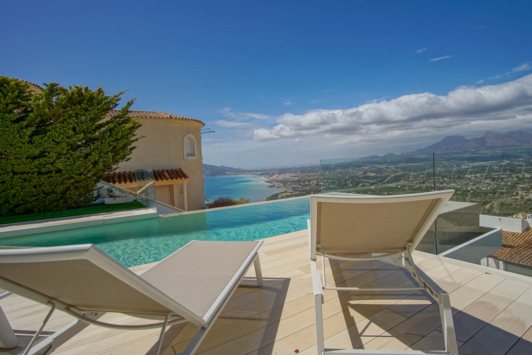 La terrasse extérieure comprend une piscine à débordement offrant une vue sur le littoral et les montagnes lointaines. Deux chaises longues sur un pont clair, bordées d'une balustrade en verre et d'une façade de villa.