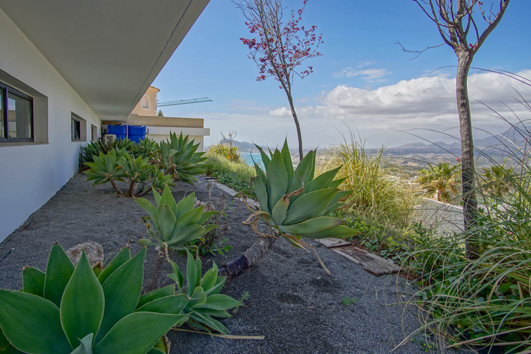 Le jardin latéral présente de grands agaves sur gravier foncé, bordant une façade blanche avec fenêtres sombres. Il offre des vues panoramiques sur la mer et les montagnes lointaines.